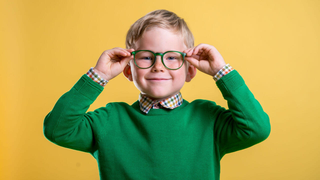 Retrato de menino loiro de suéter verde e óculos. Criança segurando óculos e olhando para a câmera.