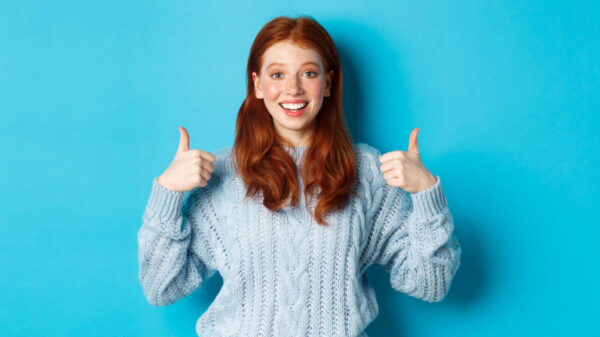 Adolescente alegre com cabelo ruivo, mostrando os polegares, de pé sobre fundo azul.