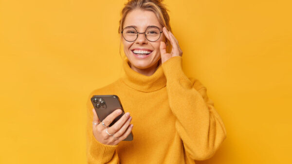 Mulher positiva sorrindo alegremente, mantendo a mão na borda dos óculos, sentindo-se alegre, usando um suéter confortável, estando contra um fundo amarelo.
