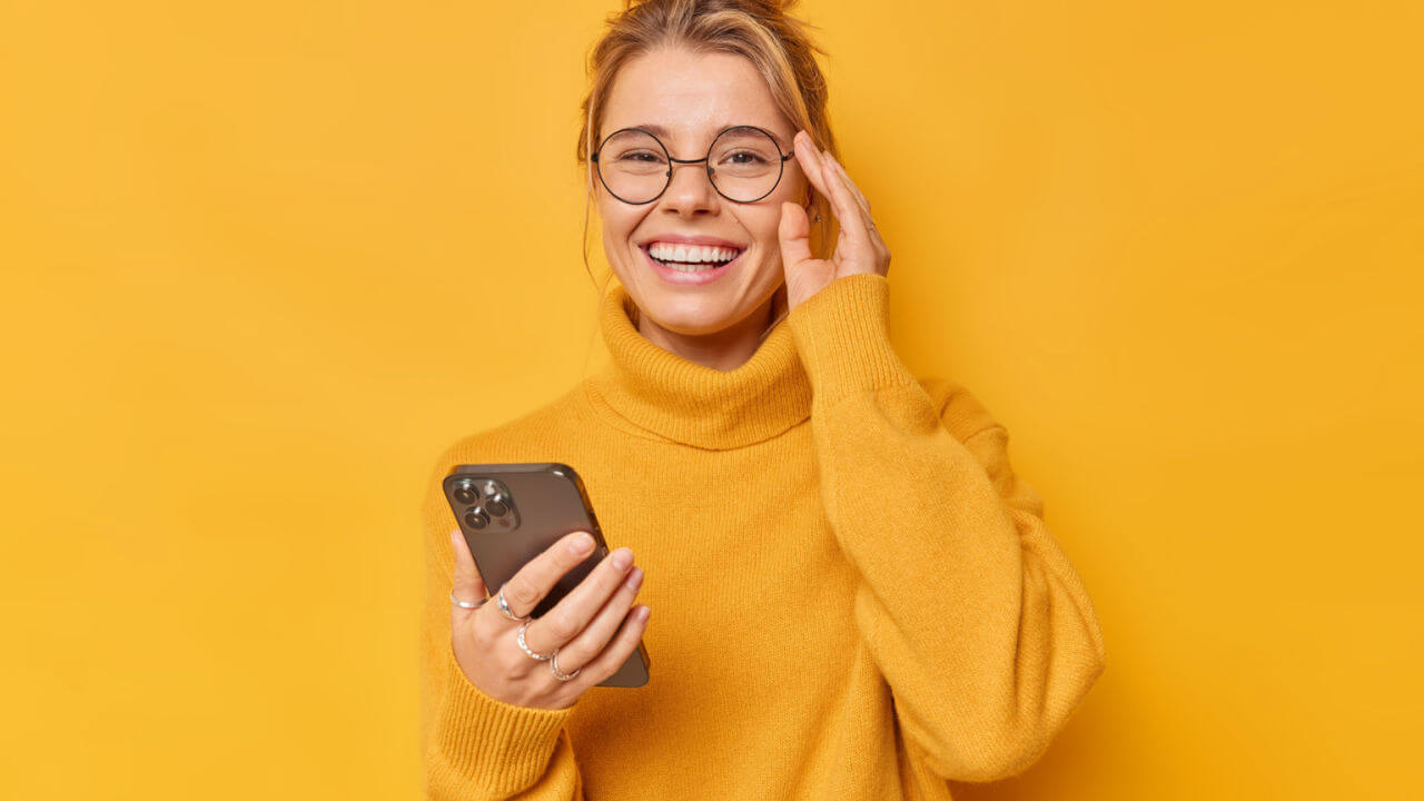 Mulher positiva sorrindo alegremente, mantendo a mão na borda dos óculos, sentindo-se alegre, usando um suéter confortável, estando contra um fundo amarelo.