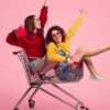 Vista lateral de jovens amigas animadas, sorrindo e levantando as mãos enquanto estão dentro de carrinho de compras, contra um fundo rosa.