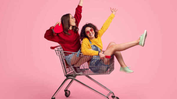 Vista lateral de jovens amigas animadas, sorrindo e levantando as mãos enquanto estão dentro de carrinho de compras, contra um fundo rosa.