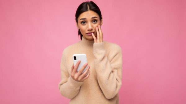 Mulher jovem chateada, triste, morena, vestindo suéter bege, isolada sobre fundo rosa, segurando celular, olhando para a câmera.