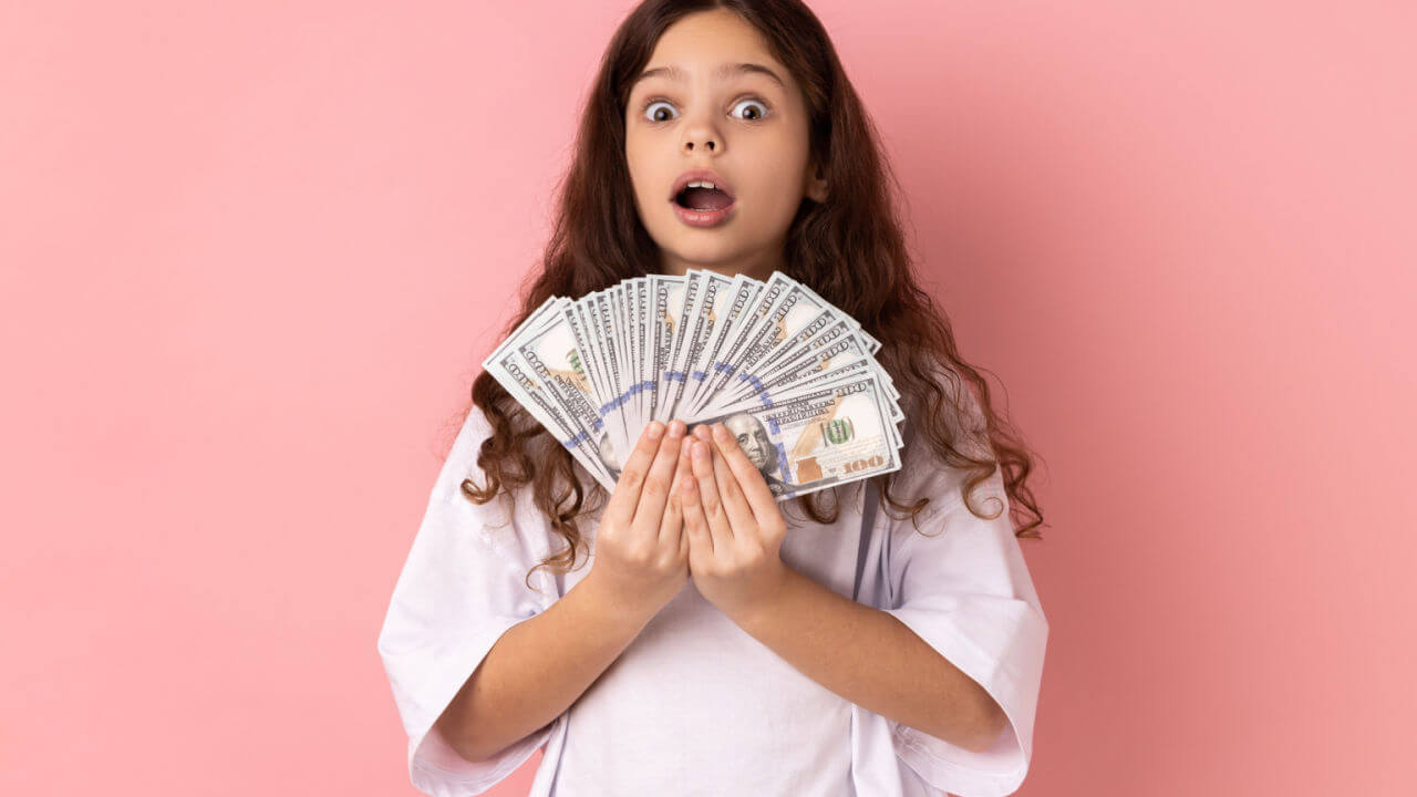 Retrato de menina rica, chocada, vestindo camiseta branca, segurando notas de dólar, olhando para a câmera com expressão de surpresa. Foto de estúdio interno isolado em fundo rosa.