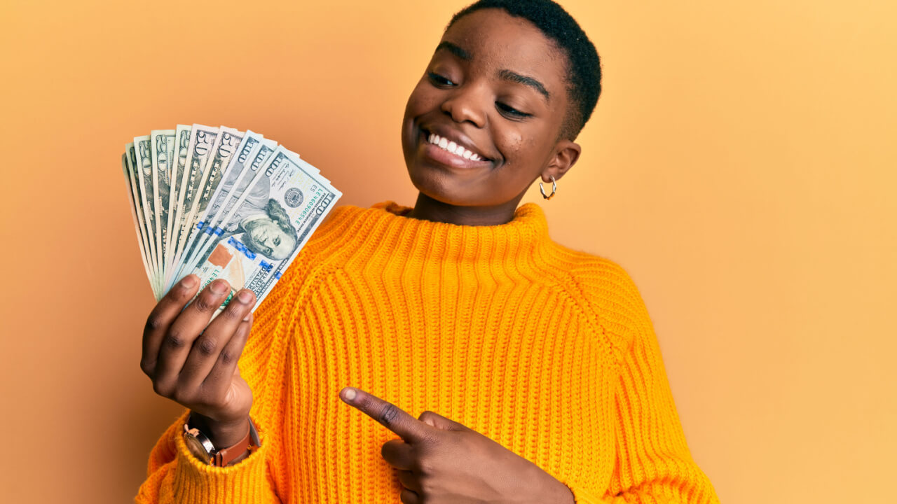 Jovem mulher afro-americana segurando dólares sorrindo feliz apontando com a mão e o dedo