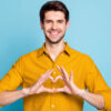 Foto de um homem sorridente fazendo um sinal em forma de coração, isolado sobre fundo de cor pastel azul.