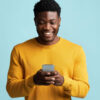 Homem segurando smartphone moderno, olhando para a tela do celular, sorrindo, em um fundo azul de um estúdio.