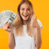 Retrato de uma jovem loira sorrindo, mostrando um monte de notas de dinheiro, com o polegar para cima, isolada sobre fundo amarelo.