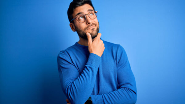 Jovem com barba vestindo suéter casual e usando óculos, sobre fundo azul. Pensando e refletindo sobre uma pergunta, com a mão no queixo.