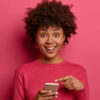 Mulher positiva, apontando para smartphone, feliz, usando blusa rosa, impressionada com algo.