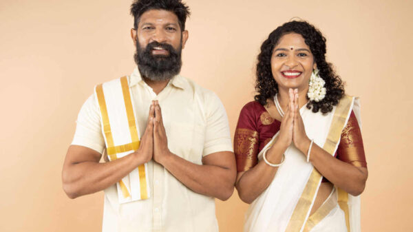 Casal feliz da Índia usando roupa branca tradicional, em pé, em uma pose de saudação com as mãos, "Namastê", isolados em fundo bege.