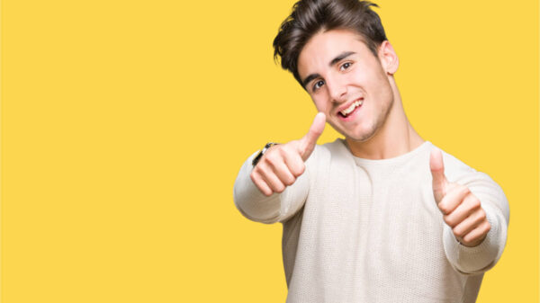 Homem sorrindo, fazendo sinal positivo com os polegares, usando uma blusa branca de manga comprida, sobre fundo amarelo.