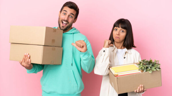 Jovem casal se mudando, segurando caixas, isolados em fundo rosa.