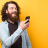 Foto de homem barbudo usando celular em um fundo amarelo.
