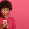 Retrato de uma garota de cabelos encaracolados olhando para o lado positivamente, usando smartphone moderno, usando suéter rosa, posando no interior de um estúdio.