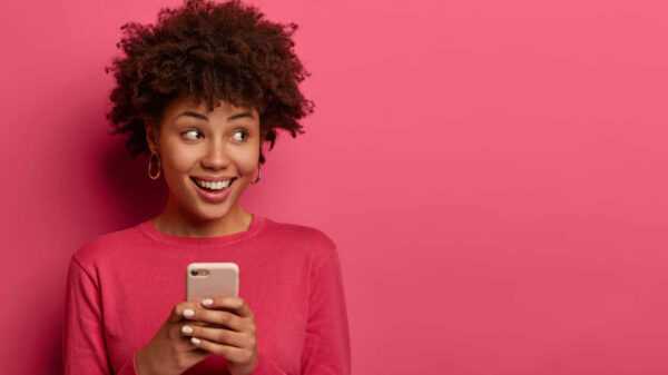 Retrato de uma garota de cabelos encaracolados olhando para o lado positivamente, usando smartphone moderno, usando suéter rosa, posando no interior de um estúdio.