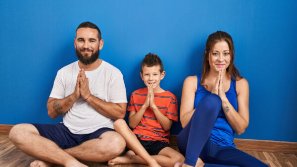 Família de três pessoas sentada no chão, em casa, orando com as mãos juntas, sorrindo confiantemente.