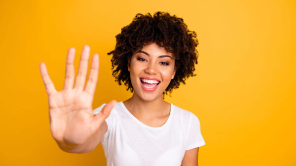 Foto de mulher feliz, com uma mão mostrando os cinco dedos levantados, vestindo uma camiseta branca casual, isolada em um fundo amarelo.