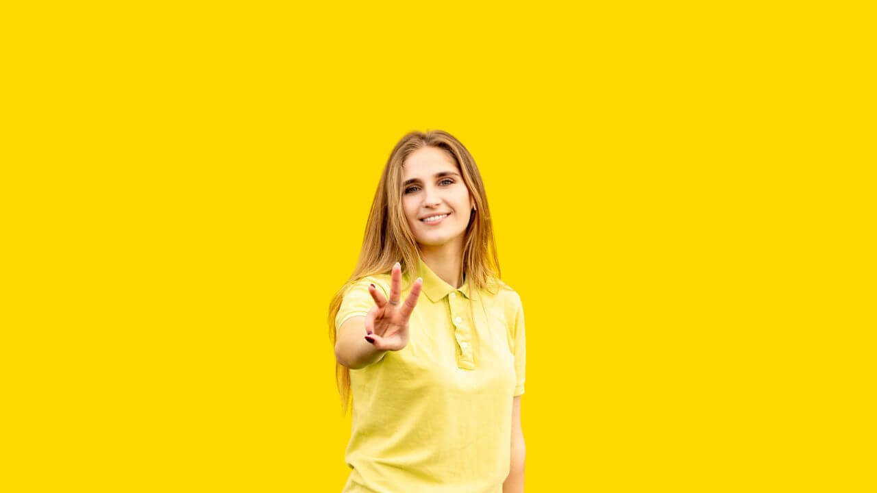 Jovem mostrando três dedos, vestida com uma camiseta amarela. Isolada no fundo amarelo.