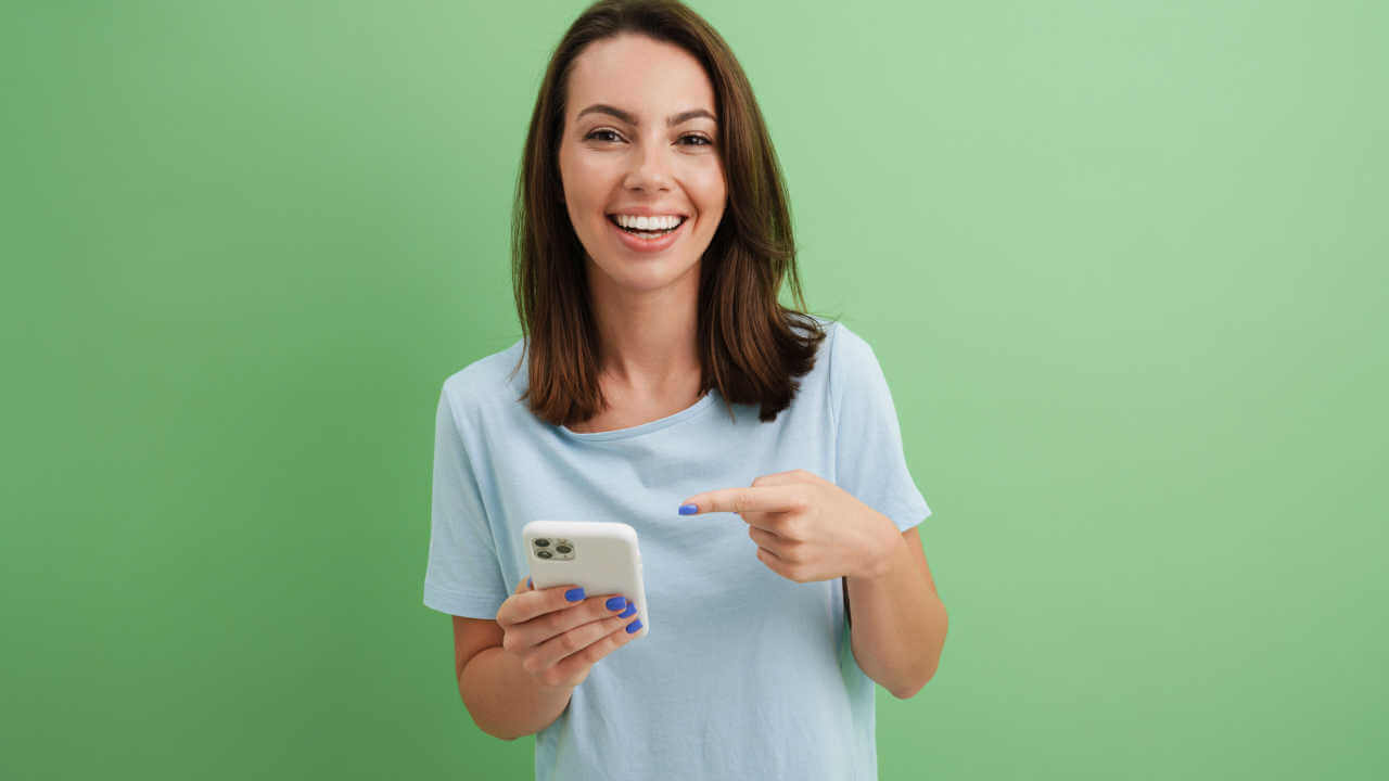 Jovem mulher rindo enquanto aponta o dedo para o celular dela, isolada sobre fundo verde.