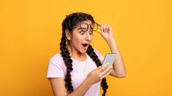 Retrato de mulher surpresa, animada, segurando e usando Smartphone, navegando nas mídias sociais. Moça emocional tirando os óculos, em fundo amarelo de um estúdio.