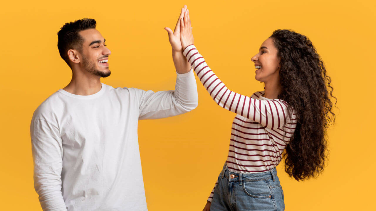 Retrato de um jovem casal feliz, fazendo "high five", alegres, em pé sobre um fundo amarelo de um estúdio.