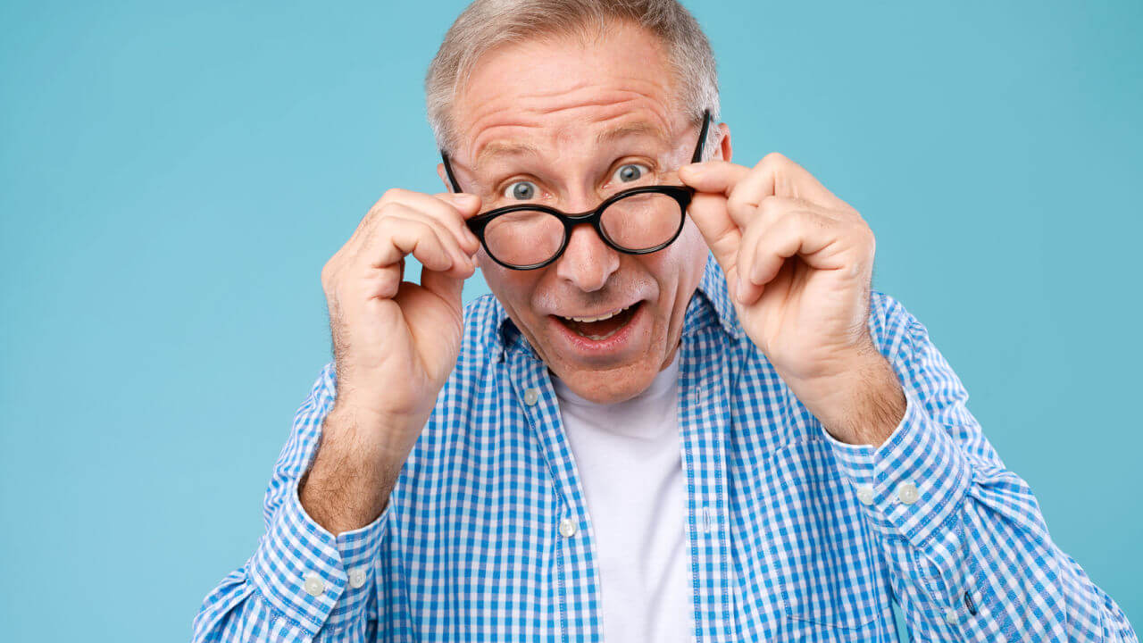 Retrato de um homem idoso olhando para a câmera, tirando ou colocando óculos, isolado em um fundo azul de um estúdio.