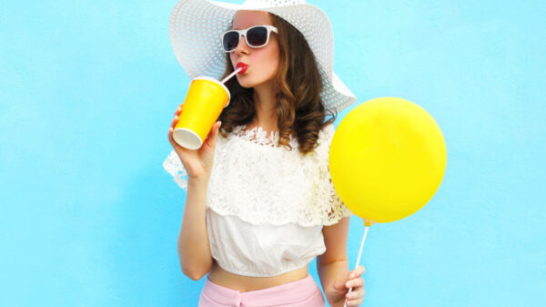 Jovem usando um chapéu de praia e um óculos de sol, segurando um balão de ar e um copo, sobre um fundo azul.