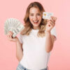 mulher com roupa branca segurando dinheiro e cartão de crédito em fundo rosa
