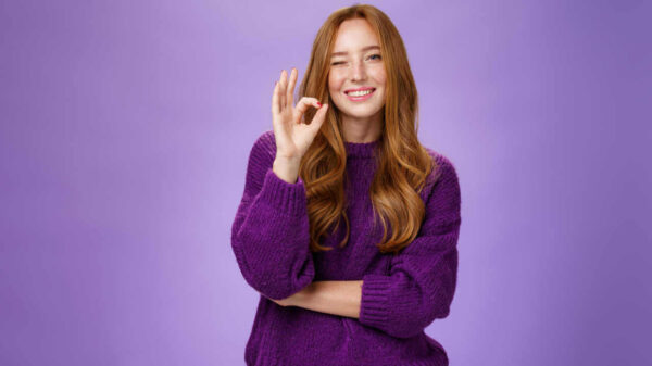 Mulher ruiva satisfeita e feliz, de suéter roxo, sorrindo e piscando em sinal de aprovação, fazendo gesto de aprovação, gostando de algo, sobre uma parede violeta.