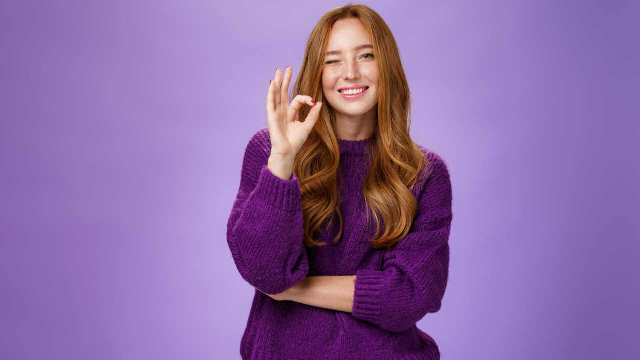 Mulher ruiva satisfeita e feliz, de suéter roxo, sorrindo e piscando em sinal de aprovação, fazendo gesto de aprovação, gostando de algo, sobre uma parede violeta.