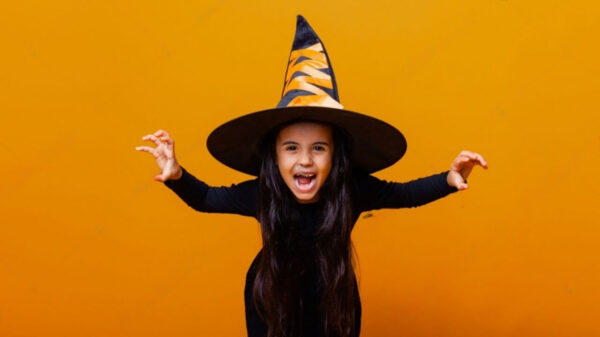Retrato de uma menina usando uma fantasia de bruxa, sobre um fundo laranja.