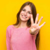 Mulher jovem, com cabelo comprido, sobre parede amarela isolada, feliz e fazendo o número quatro com os dedos.