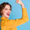 Foto de uma mulher muito alegre, mostrando o bíceps. Mulher forte e poderosa, usando camisa amarela, isolada em um fundo azul.