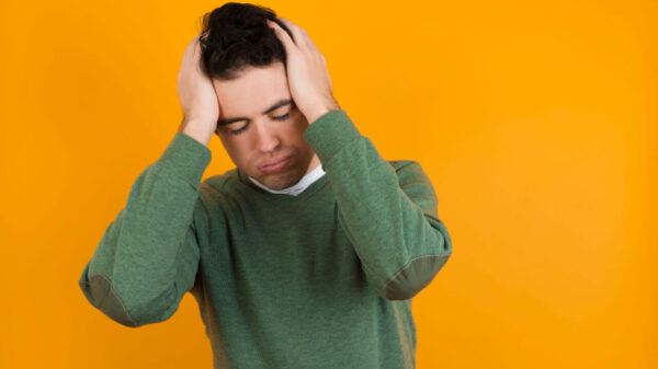 Homem jovem vestindo suéter verde contra uma parede laranja, sofrendo, desesperado e estressado. Conceito de paranoia.