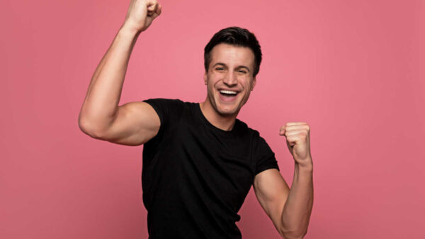 Foto de um jovem usando uma camiseta preta, com os punhos fechados, comemorando e sorrindo.
