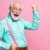 Foto de idoso levantando os punhos, celebrando, com bom humor, usando camisa de cor menta, suspensórios, gravata borboleta violeta e calça, isolado em um fundo de cor rosa pastel.