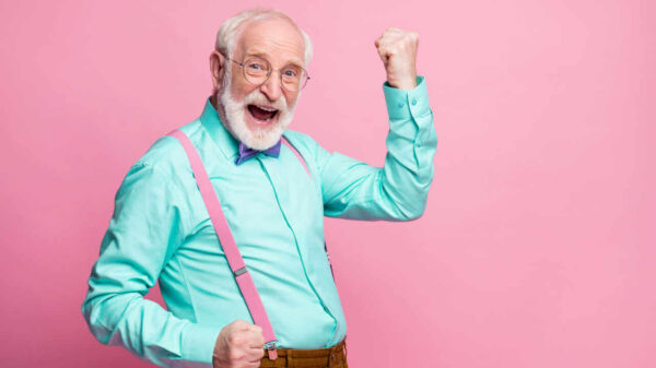 Foto de idoso levantando os punhos, celebrando, com bom humor, usando camisa de cor menta, suspensórios, gravata borboleta violeta e calça, isolado em um fundo de cor rosa pastel.