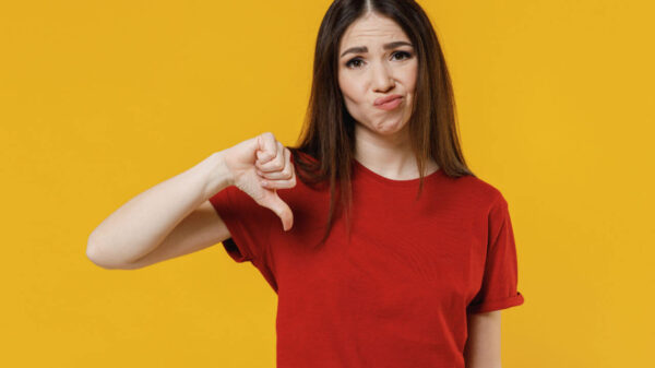 Mulher insatisfeita morena, usando camiseta vermelha básica, mostrando o polegar para baixo em gesto de negatividade, isolada em um estúdio de fundo amarelo.