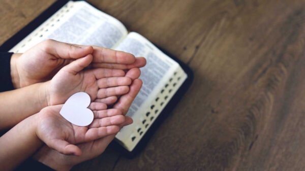 Mãos segurando um coração de papel branco em formato de coração. Bíblia em segundo plano.
