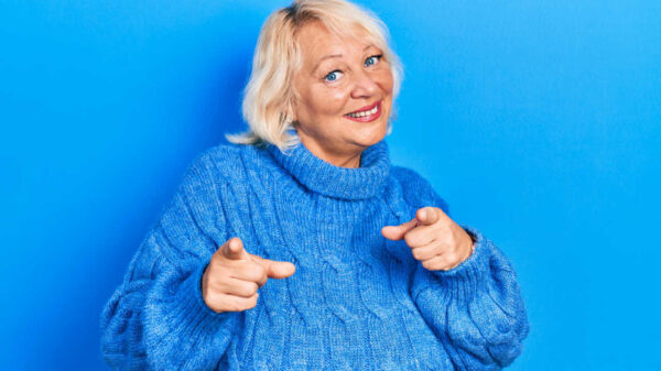 Mulher loira de meia-idade vestindo roupas casuais, apontando os dedos para a câmera, com cara feliz.