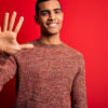 Jovem vestindo suéter casual, em pé, sobre fundo vermelho, mostrando os dedos fazendo o número cinco enquanto sorri confiante e feliz.