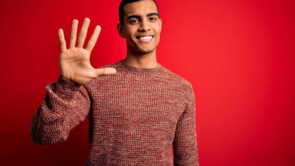 Jovem vestindo suéter casual, em pé, sobre fundo vermelho, mostrando os dedos fazendo o número cinco enquanto sorri confiante e feliz.