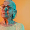 Mulher idosa inteligente, usando óculos redondos, olhando para a câmera. Senhora sábia com cabelo penteado para trás, isolada sobre fundo laranja.