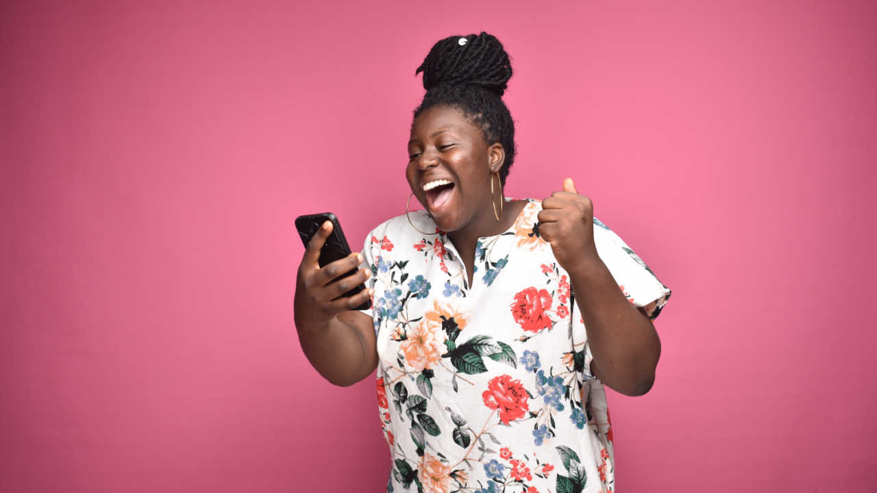 mulher negra celebrando com o celular na mão em fundo rosa