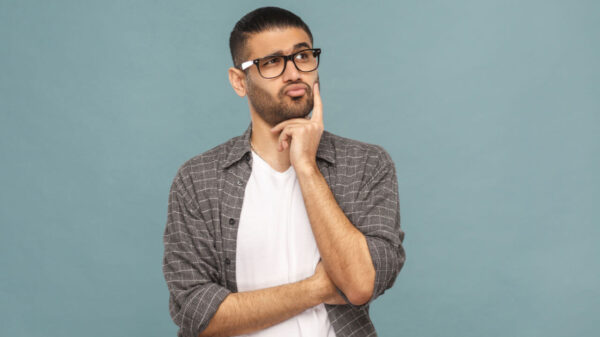 Retrato de homem pensativo, barbudo, com óculos pretos, em estilo casual, pensando. Estúdio com fundo azul.