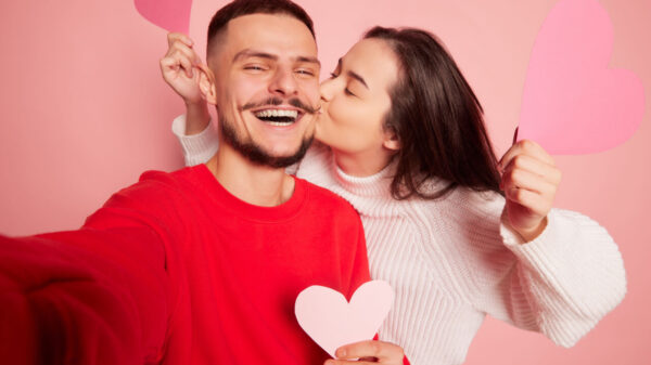 Retrato de um casal jovem. Mulher beijando homem feliz e sorridente tirando selfie dos dois juntos. Isolados sobre fundo rosa. Conceito de amor, relacionamento, emoções e estilo de vida.