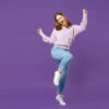 Jovem feliz. Foto de corpo inteiro. Mulher vestindo suéter lilás, apontando os dedos para cima, com uma perna levantada, isolada em um fundo violeta. Retrato de estúdio Conceito de estilo de vida.