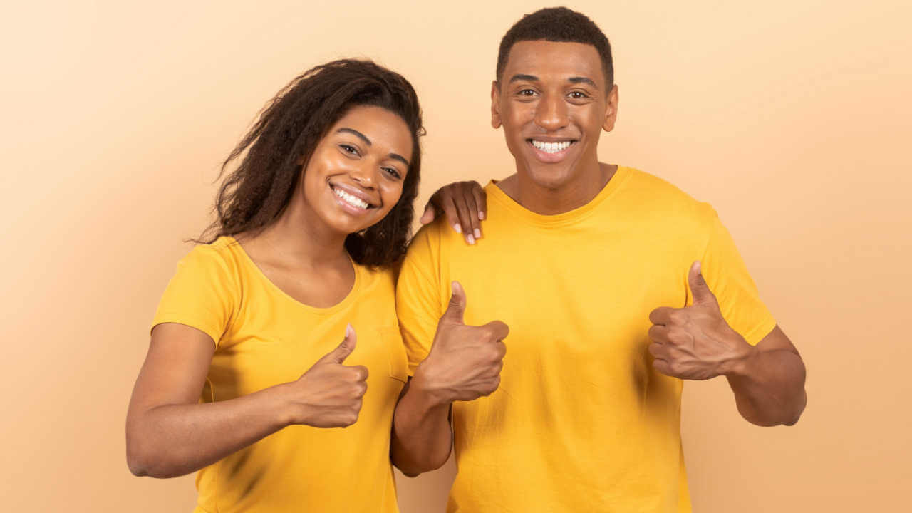 Casal feliz, mostrando polegares para cima, fazendo gesto de aprovação. Homem e mulher posando sobre fundo de um estúdio cor-de-pêssego.