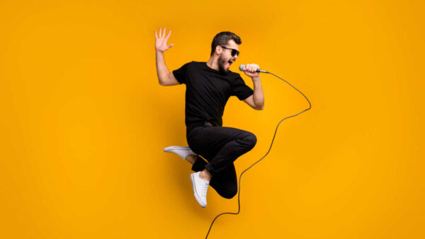 homem de preto pulando com microfone na mão em fundo amarelo